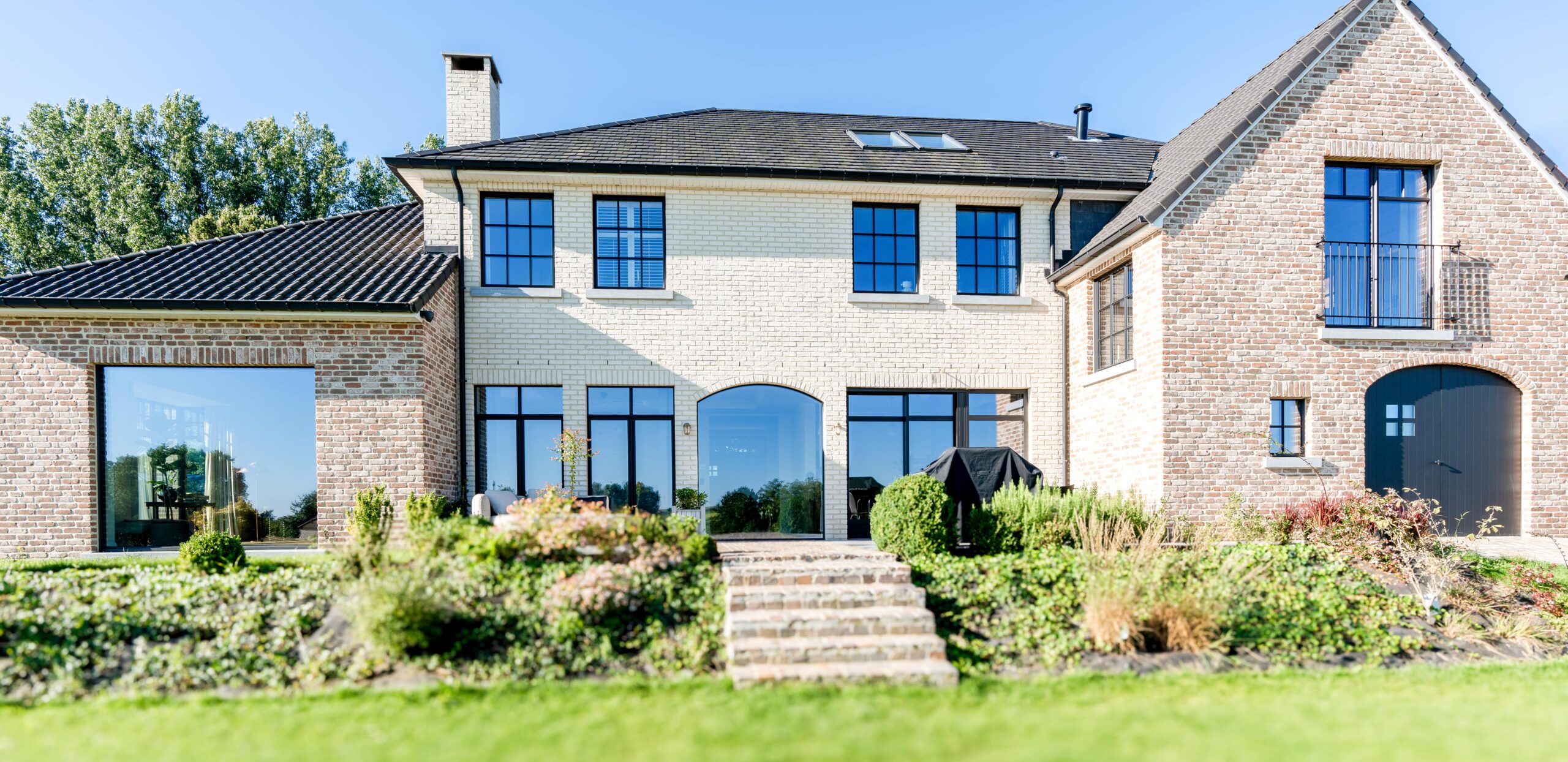 Une grande maison en brique à deux étages avec un toit noir, de multiples fenêtres et une cheminée. La cour avant comprend un jardin soigneusement aménagé avec des marches menant à l'entrée. Le ciel est clair et bleu.