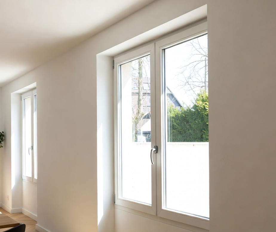 Une pièce moderne et lumineuse avec de grandes fenêtres à cadre blanc qui laissent entrer la lumière naturelle. À l'extérieur, on aperçoit des arbres et une partie d'un bâtiment. Les murs sont blancs, ce qui crée une atmosphère propre et aérée.